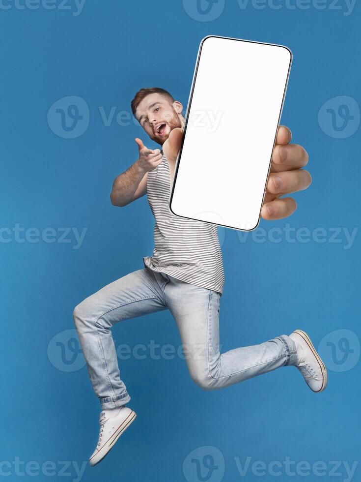 Emotional millennial redhaired guy pointing finger at camera, jumping with mobile phone on blue studio background, mockup for mobile app on white screen. Creative collage with website template photo