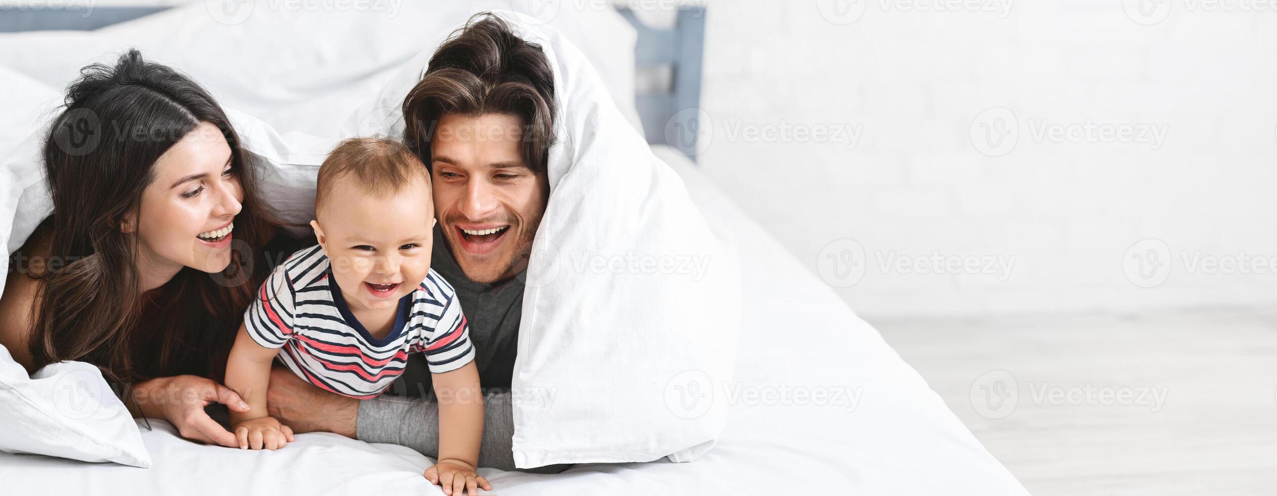 A happy family relaxes in a cozy bedroom, sharing delightful moments while playing under a fluffy blanket. The baby giggles joyfully, while parents smile at each other and their child, copy space photo