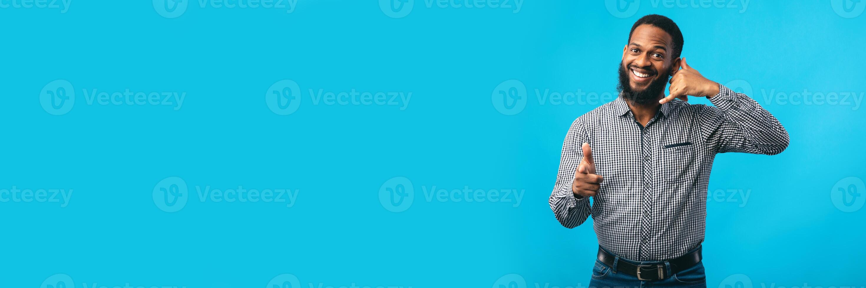 Playful african american man pointing index finger at camera, doing phone gesture saying call me back, isolated over turquoise wall, copy space, web-banner photo