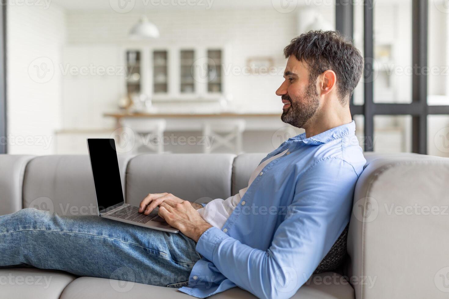 This image shows a man relaxing on a gray sofa, using a laptop. He is wearing jeans and a light blue button-up shirt and is looking at the laptop screen while typing. photo