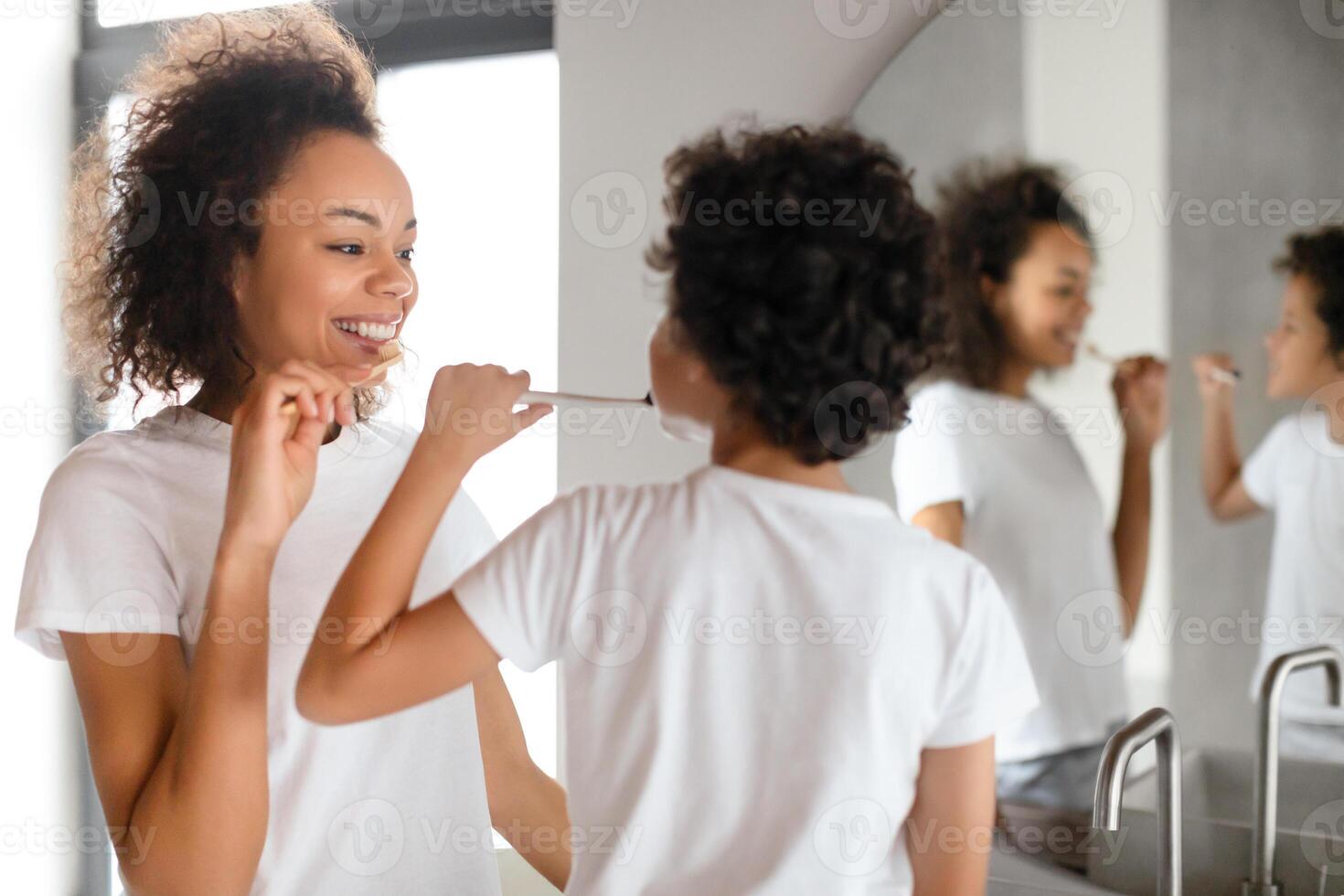 African American young woman with a bright smile is brushing her teeth in front of a bathroom mirror, dressed in a casual white t-shirt and is actively engaged in her daily oral hygiene practice. photo