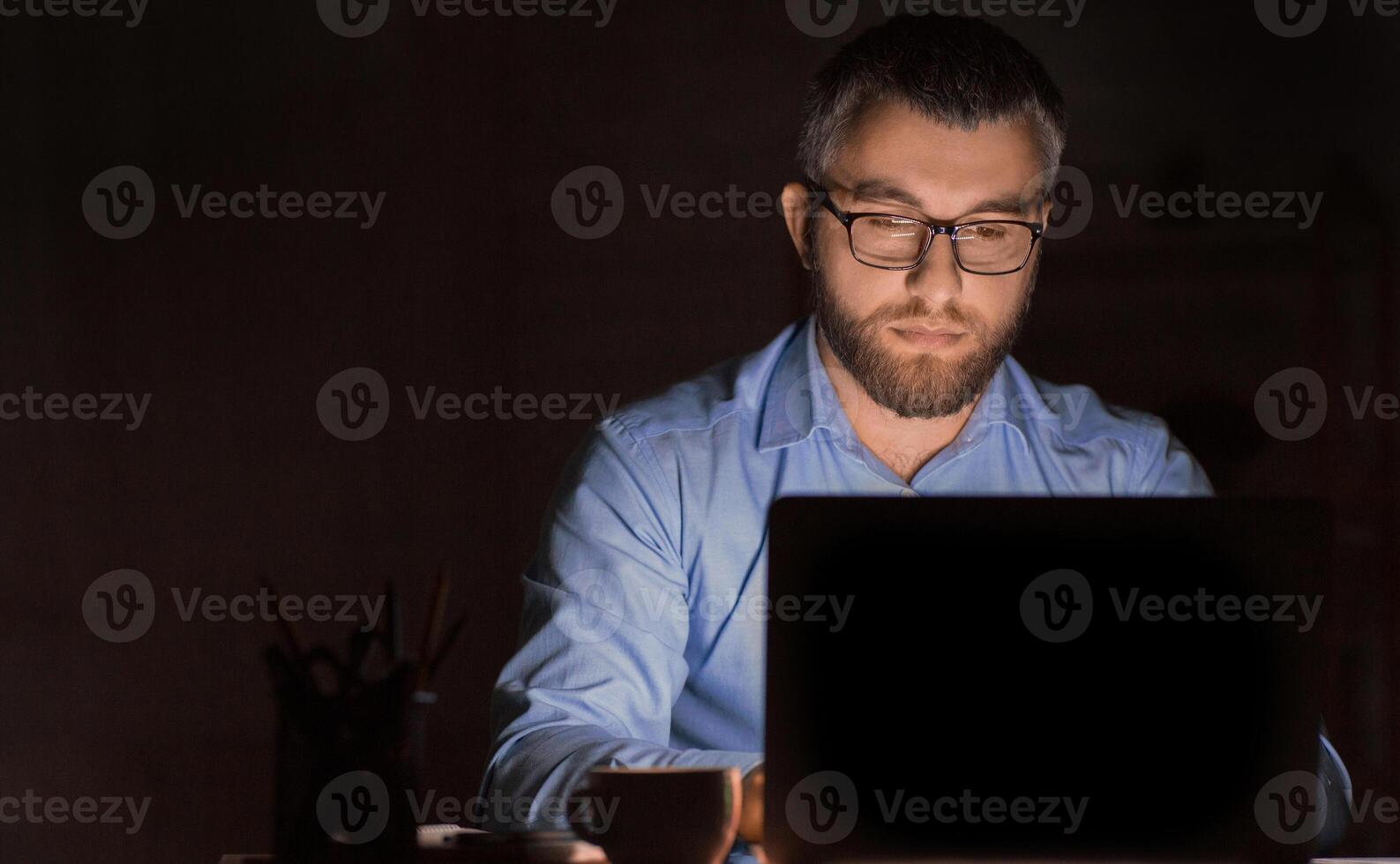 A man wearing glasses is intensely focused while working on his laptop in a dimly lit office environment during late-night hours. He appears to be deeply engaged in a task photo