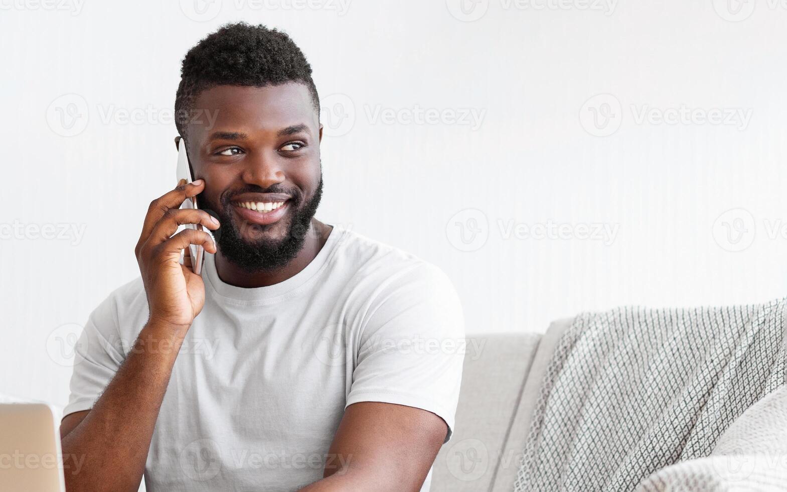 A young, African American man with a beard, wearing a white t-shirt, sits on a gray couch and smiles while talking on his phone. He is looking to the side as he speaks on the phone photo