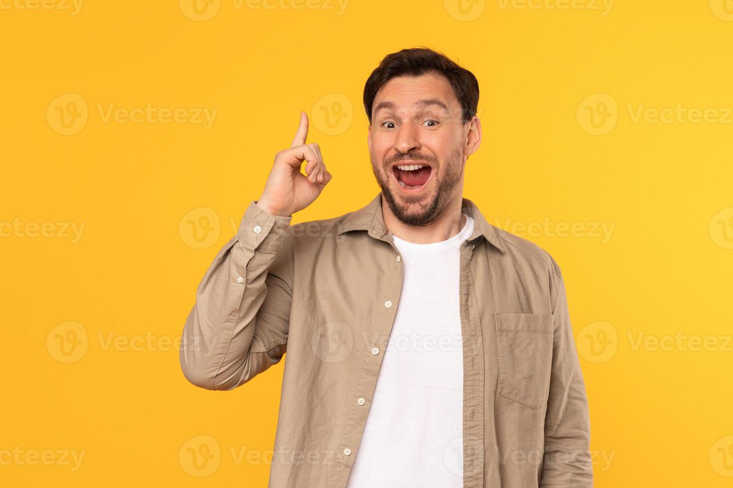 A man with a beard and short brown hair is wearing a tan shirt over a white t-shirt. He is looking at the camera with his mouth open and his index finger raised in the air photo