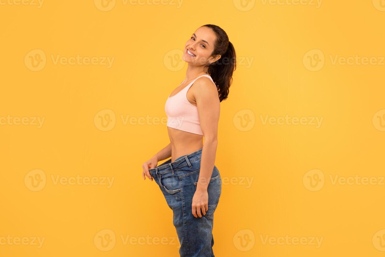 A cheerful young woman stands against yellow backdrop, holding her overly large jeans to show her successful weight loss journey. Her bright smile and confident pose convey a sense of achievement. photo