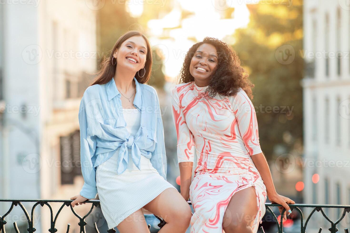 Two multiethnic young women sit on a railing in a city setting, smiling and laughing, enjoying each others company. The photo captures a moment of joy and friendship