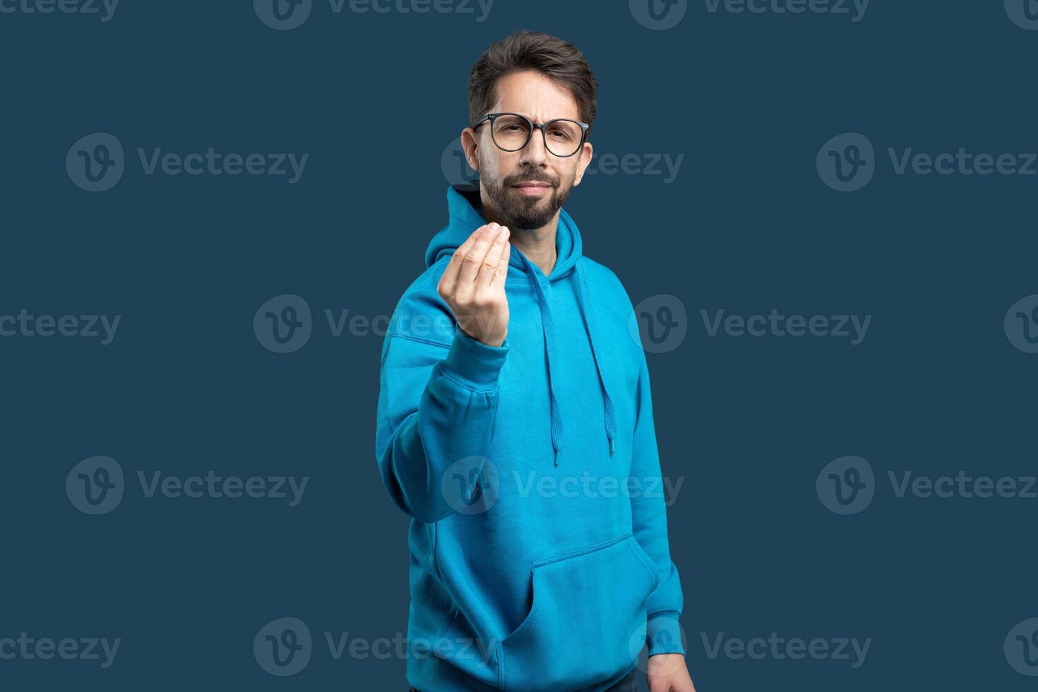 A man in a vibrant blue hoodie stands against a solid dark background, extending his hand in a gesture that suggests he is inviting or requesting something from the viewer. photo