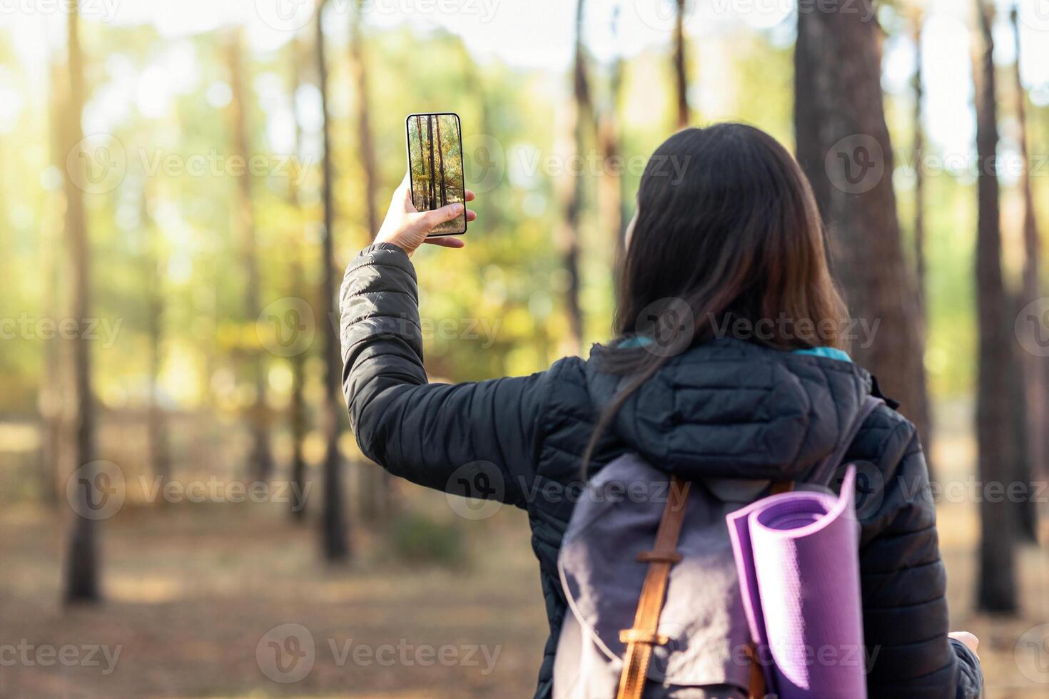 Back view of woman with backpack taking photo of forest on mobile phone, copy space