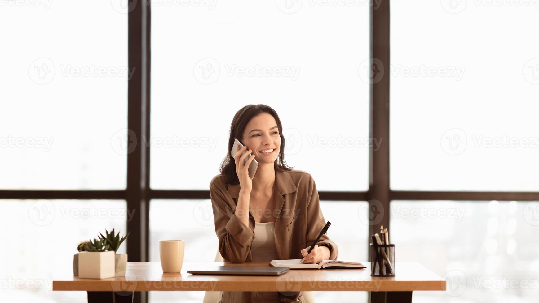 Checking Schedule. Portrait of female secretary making phone call sitting at desk, writing in her notebook, copyspace photo