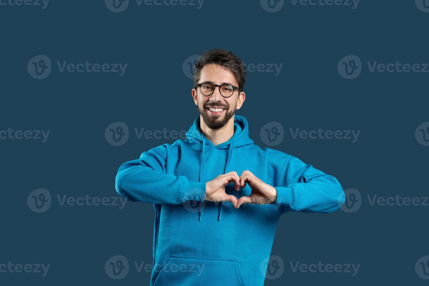 A cheerful young man wearing a blue hoodie smiles broadly while forming a heart shape with his hands. The solid dark backdrop highlights his joyful expression and gesture of love. photo