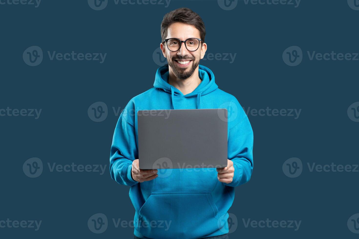 A young man wearing a bright blue hoodie smiles widely while holding a laptop in front of him. The backdrop is a simple dark color that highlights his joyful expression and tech device. photo