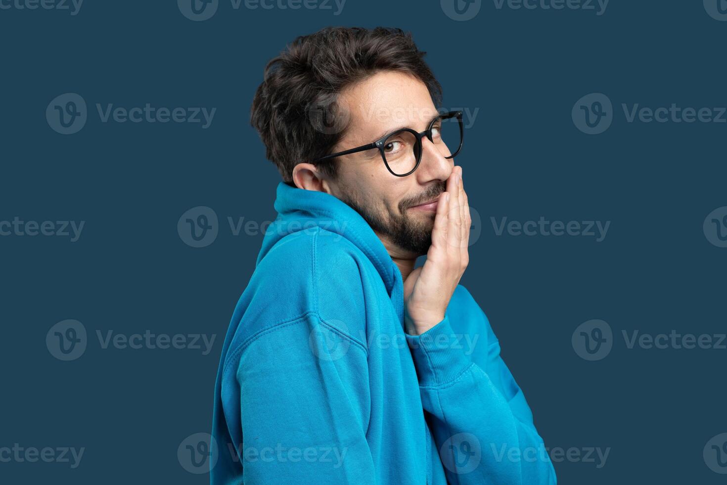 A young man is playfully hiding his face behind his hand while wearing a blue hoodie. He has thick glasses and a light-hearted expression, creating a fun indoor atmosphere. photo