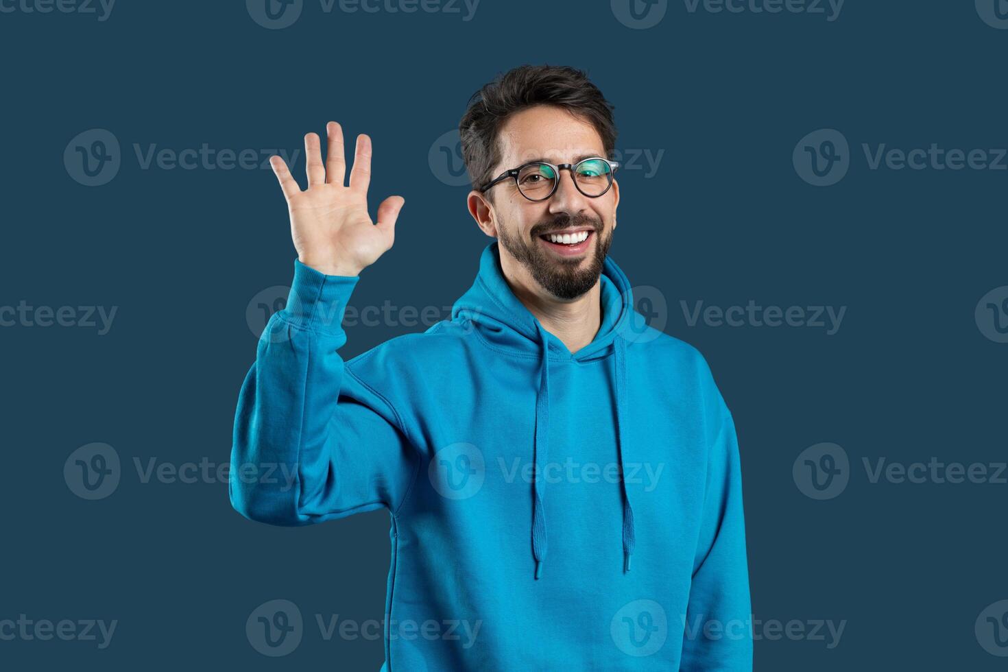 A cheerful young man greets with a smile and raised hand, dressed in a blue hoodie, set against a dark, solid backdrop. His happy expression conveys friendliness and warmth. photo