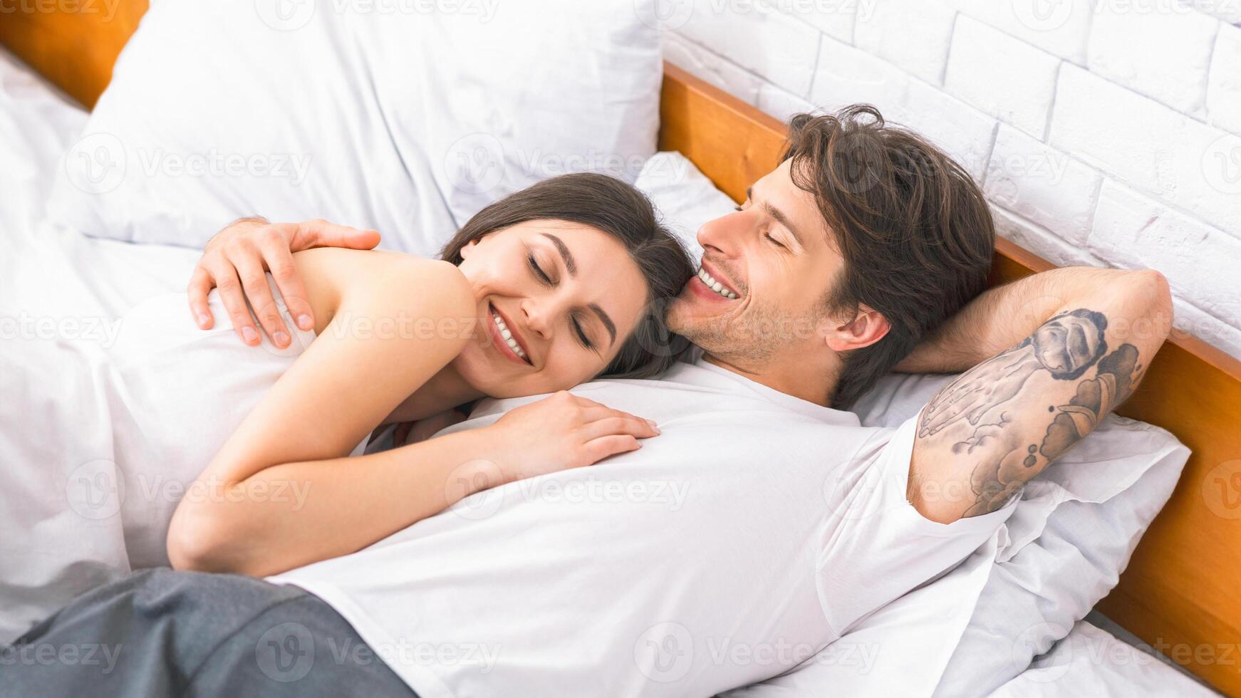 A young couple is lying in bed together, both smiling and seemingly enjoying their time together. The woman is nestled into the mans side, her head resting on his chest, and he has his arm around her. photo