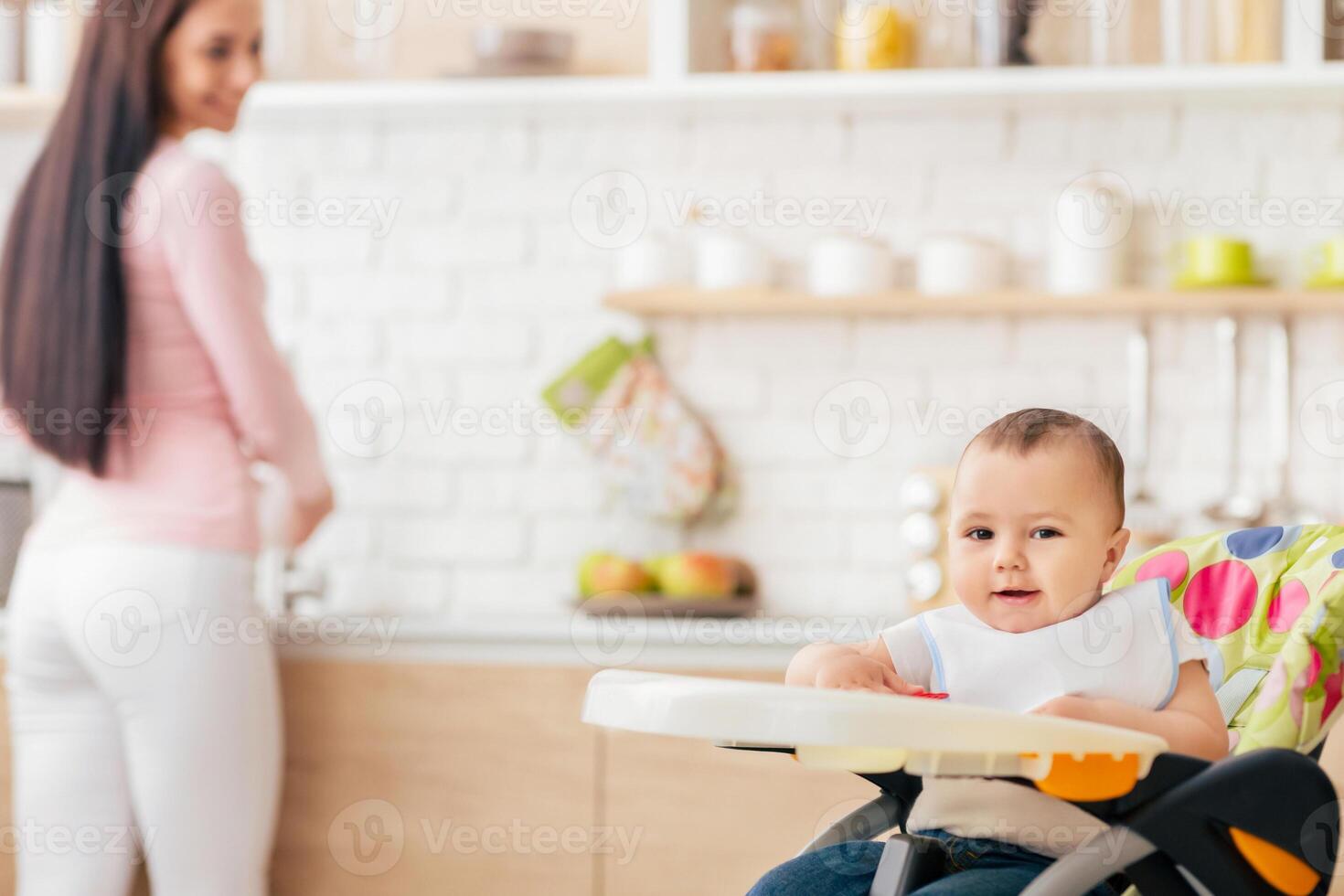 A woman is standing next to a baby seated in a high chair. The woman appears to be observing or caring for the baby. The high chair is clean and modern in design, with safety straps visible. photo