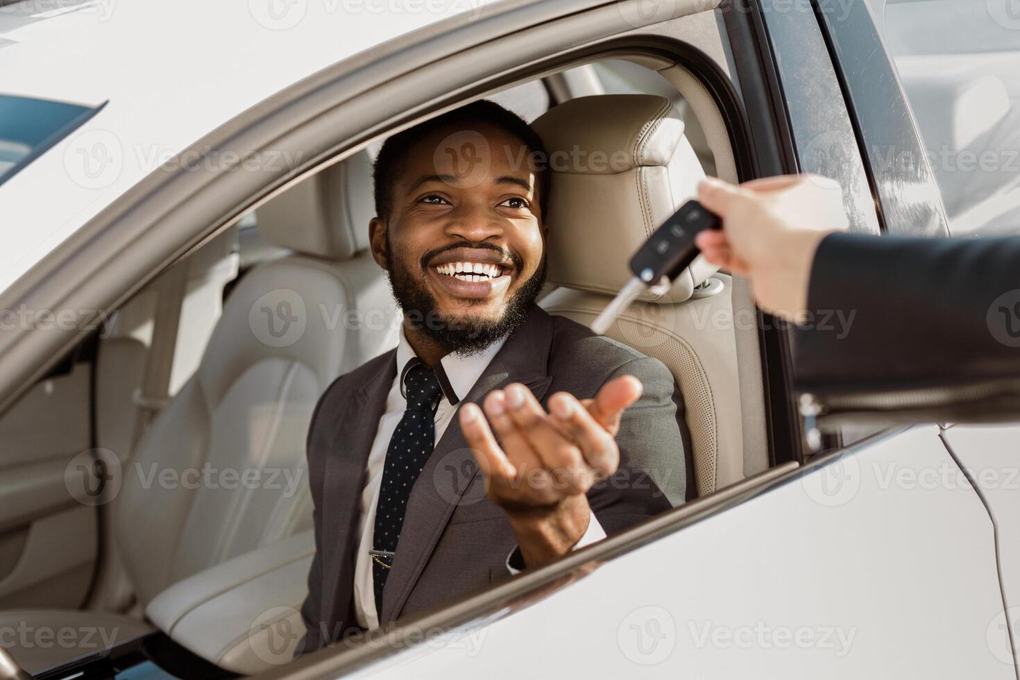 African American man in a suit sits in the drivers seat of a white car, smiling broadly as he receives the car keys from a dealer standing outside. The car is parked in a dealership lot. photo