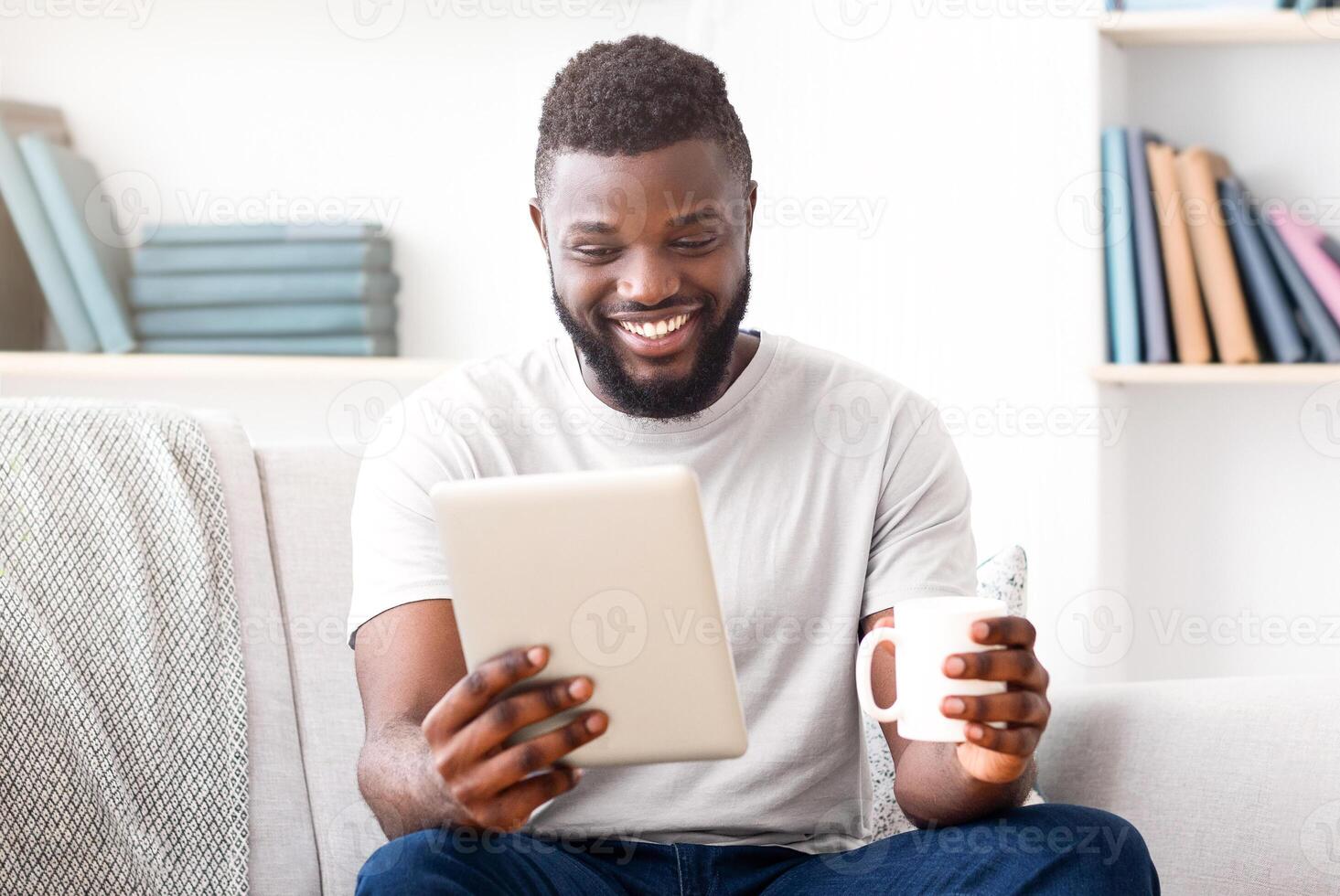 A smiling African American man wearing a white t-shirt sits on a white couch with a grey throw blanket, using a tablet and holding a white coffee cup. photo