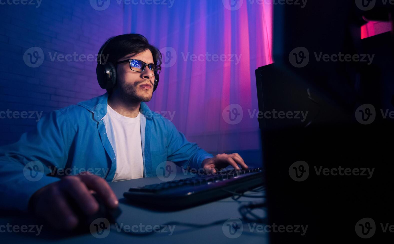 A young man sits in front of a computer, wearing headphones and blue light glasses. He is typing on the keyboard with his right hand and using a mouse with his left hand. photo