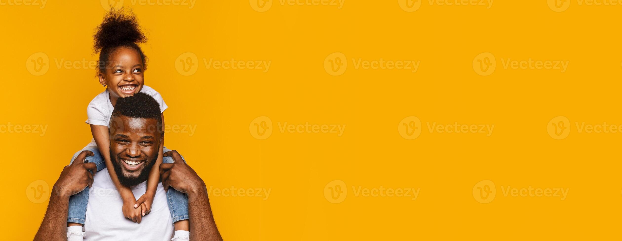 Black father is playfully giving his daughter a piggyback ride, both of them sharing bright smiles. The vibrant yellow backdrop enhances their joyful expressions, reflecting a delightful moment photo