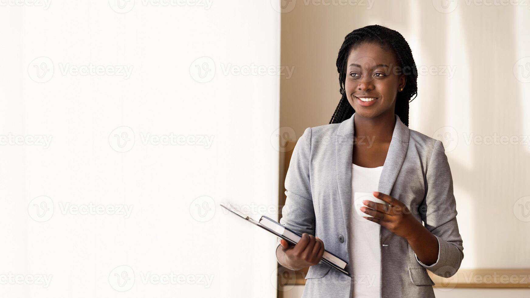 Pensive african secretary having coffee break near window in office, standing with clipboard, notebook and cup in hands, free space photo