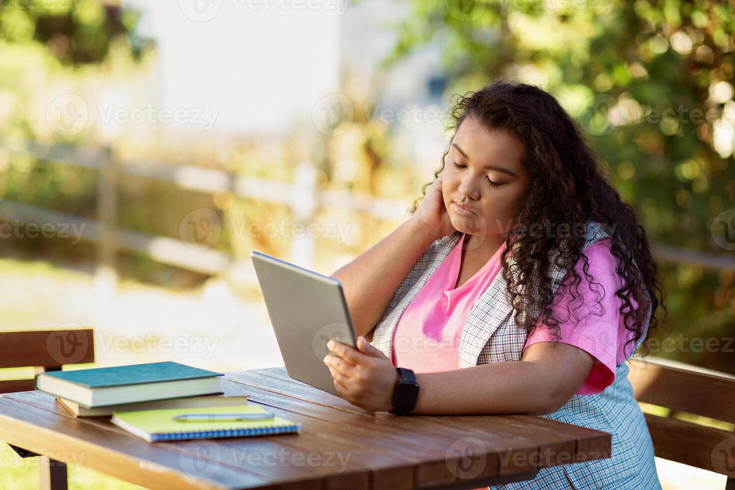 A young woman sits at a wooden table in a sunny garden, focused on her tablet. Books and notebooks are scattered nearby, suggesting a study or relaxed reading session. photo