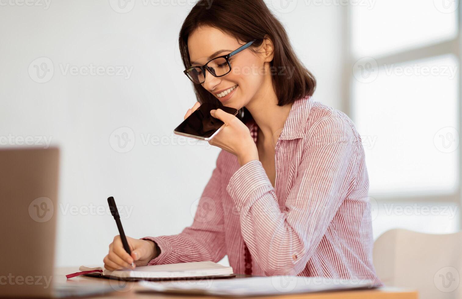 Business Communication Concept. Cheerful boss woman using voice command recorder on smartphone and taking notes photo