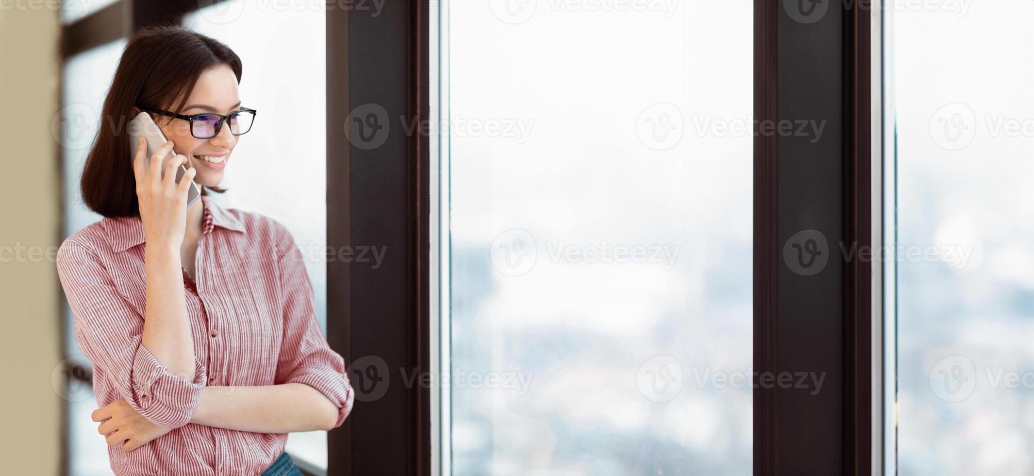 Communication Concept. Portrait of female student in specs talking on cellphone, looking out the window, copyspace photo