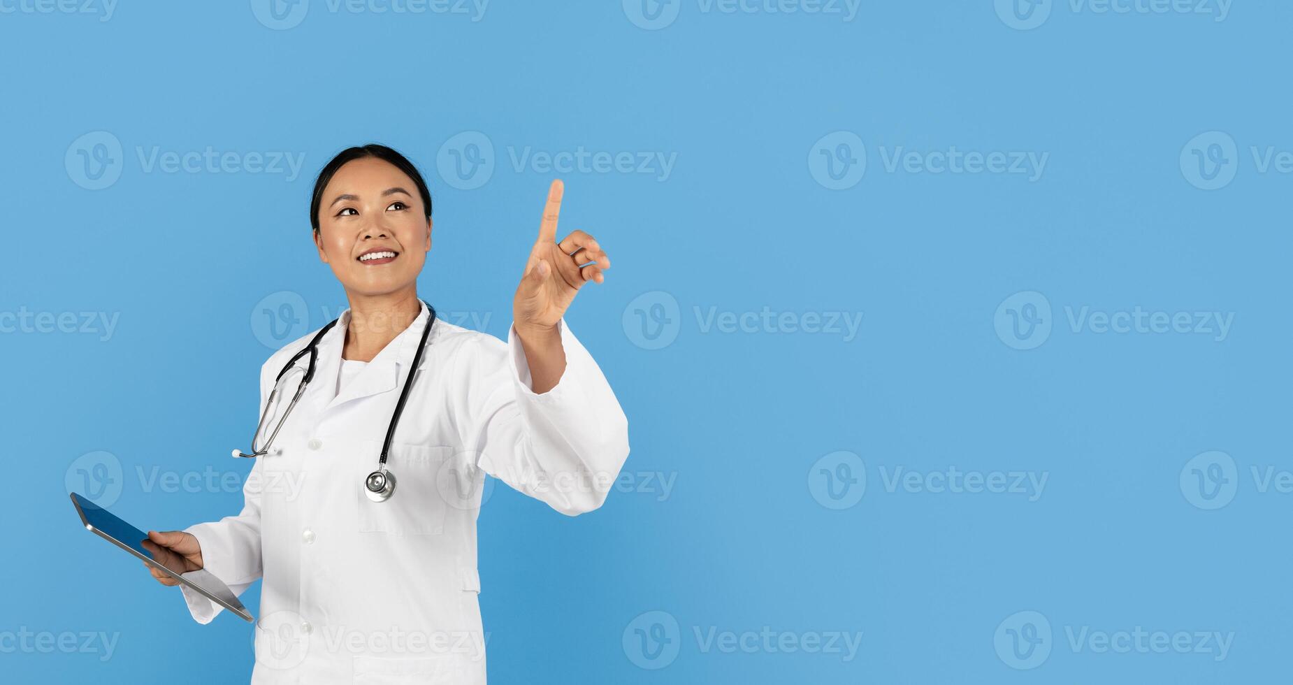A medical professional Asian lady stands confidently with a tablet, pointing upwards as if sharing important information. The background is a solid blue, enhancing the focus on her. photo