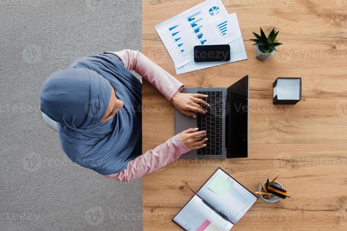 Office workspace top view. Muslim woman in hijab working on laptop with black screen at wooden desk, typing on keyboard, free space photo