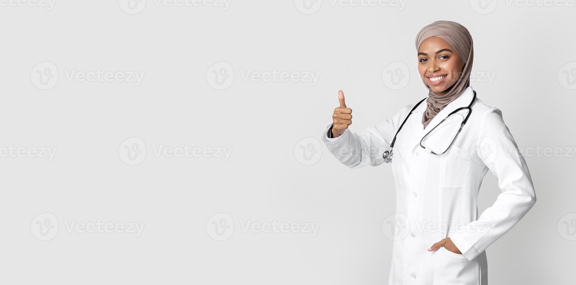 Medical Career For Muslim Women. Joyful Black Female Doctor In Headscarf And White Coat Showing Thumb Up, Posing Over Light Background, Free Space photo
