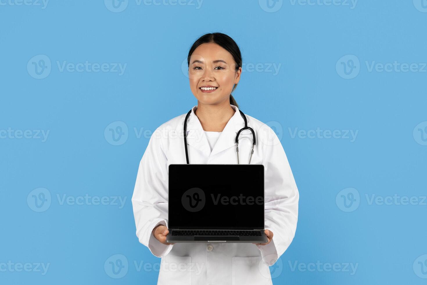 A smiling Asian woman healthcare worker in a white lab coat holds a laptop against a blue backdrop, symbolizing the integration of technology in patient care and medical practice. photo