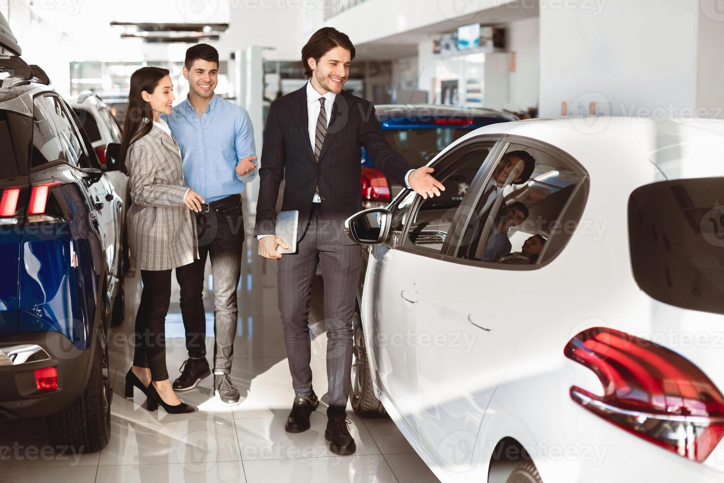 Car Rental Concept. Happy family choosing car for rent, manager showing them luxury auto standing in dealership store photo