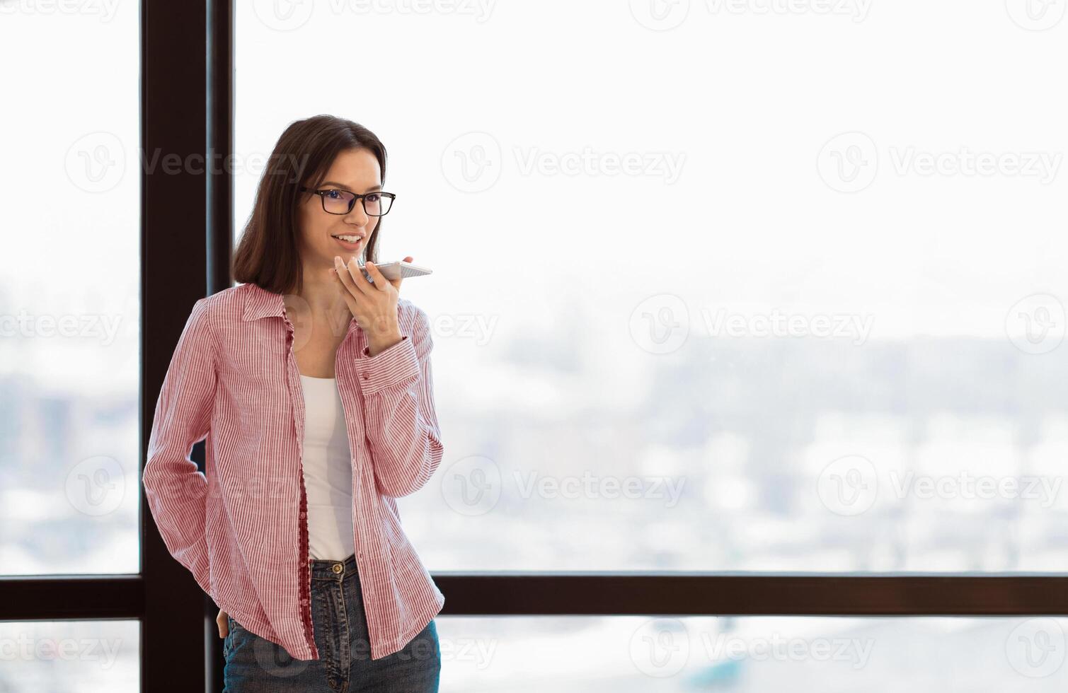 Young female manager in specs talking on speaker phone using cellphone, standing by window in office, copyspace photo