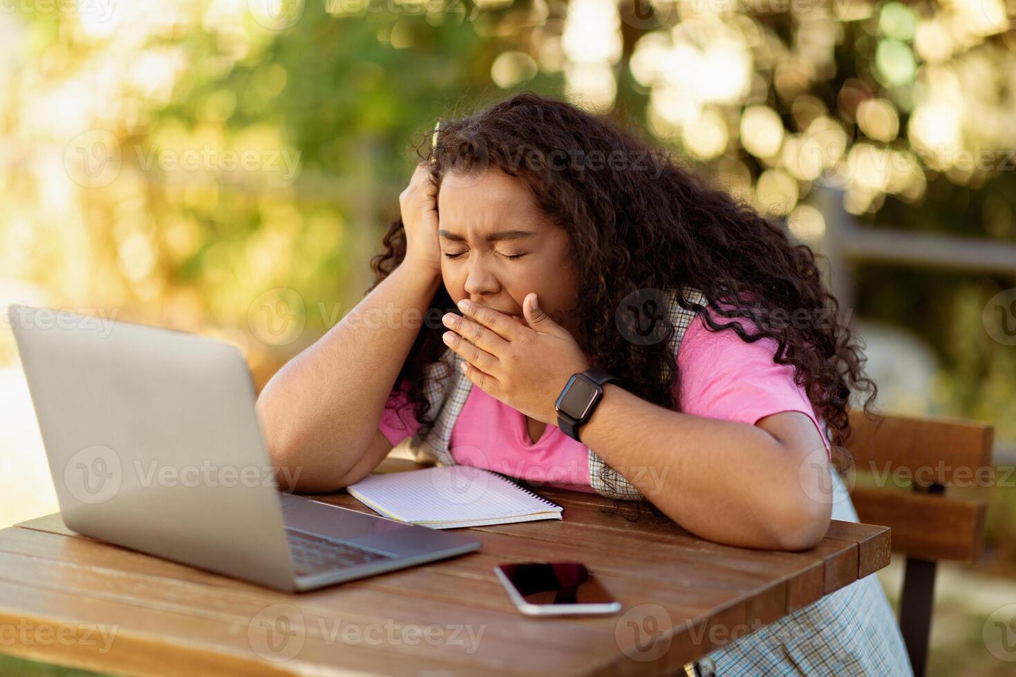 A young woman sits at a wooden table outside, working on her laptop. She appears fatigued and yawning, indicating a long study session in a sunny environment. photo