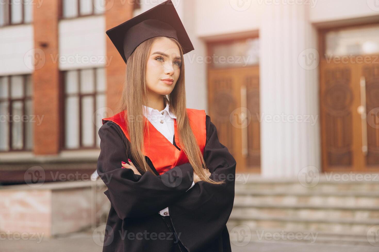 Confident Young Woman Celebrating Graduation in Cap and Gown with Arms Crossed in Front of University Building photo