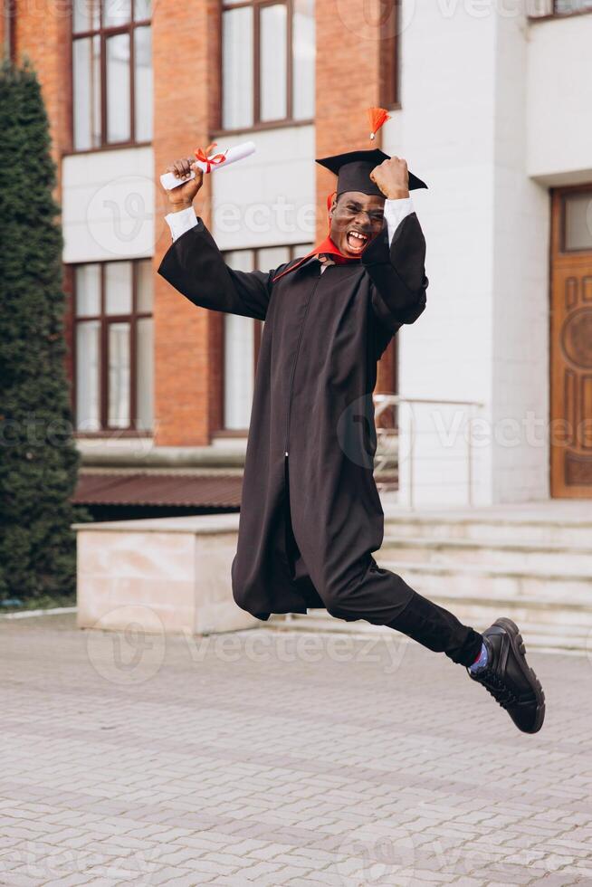 Joyful African American Male Graduate Jumping with Diploma in Hand Celebrating on Campus Outdoors photo