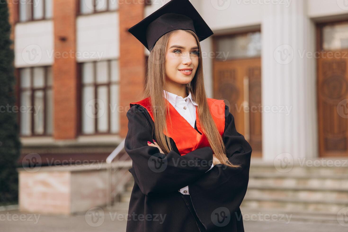 Confident Young Woman Celebrating Graduation in Cap and Gown with Arms Crossed in Front of University Building photo