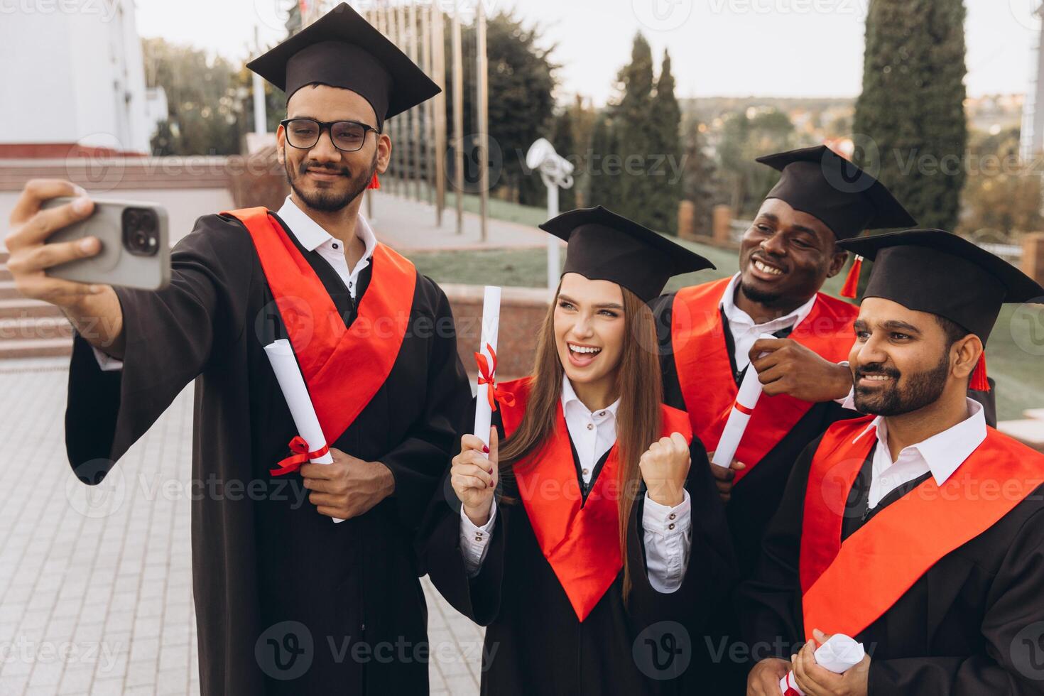 Group of Diverse Graduates Celebrating with Selfie Outdoors in Traditional Caps and Gowns photo