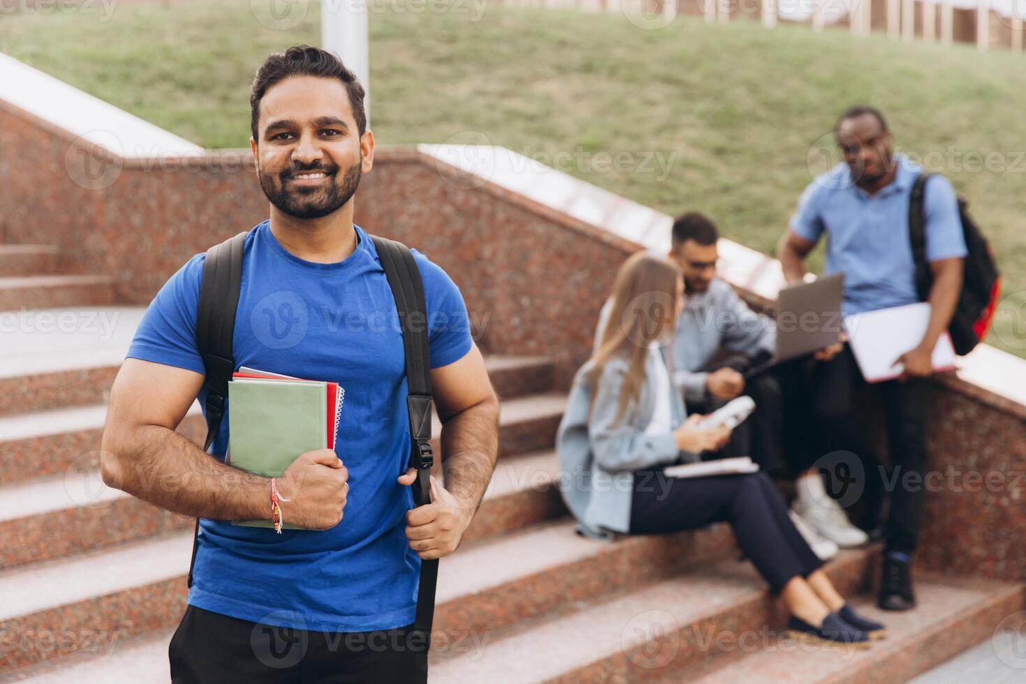 Confident Indian Student Standing with Books on Campus Steps with Group Study Session in Background photo