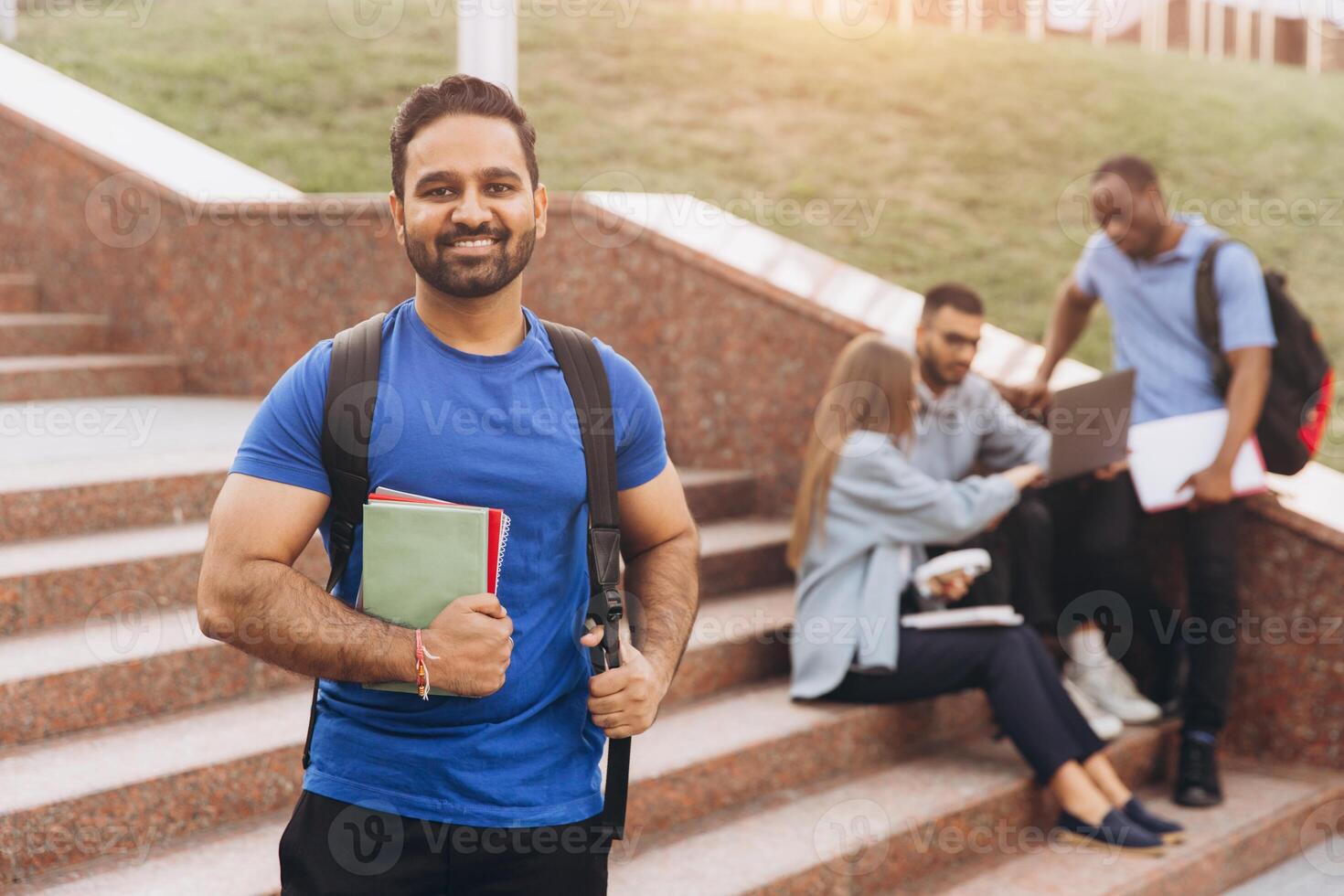 Smiling Indian Student Holding Books Outdoors with Friends Studying on Campus Steps photo