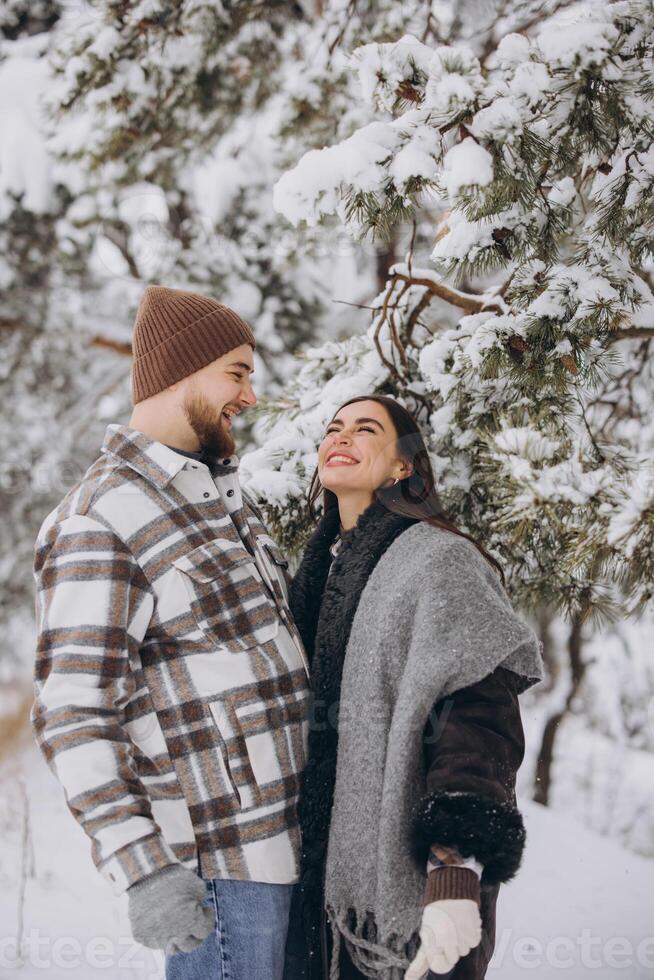 A young happy and loving couple is standing under a pine tree from which snow falls in the mountains in winter. photo
