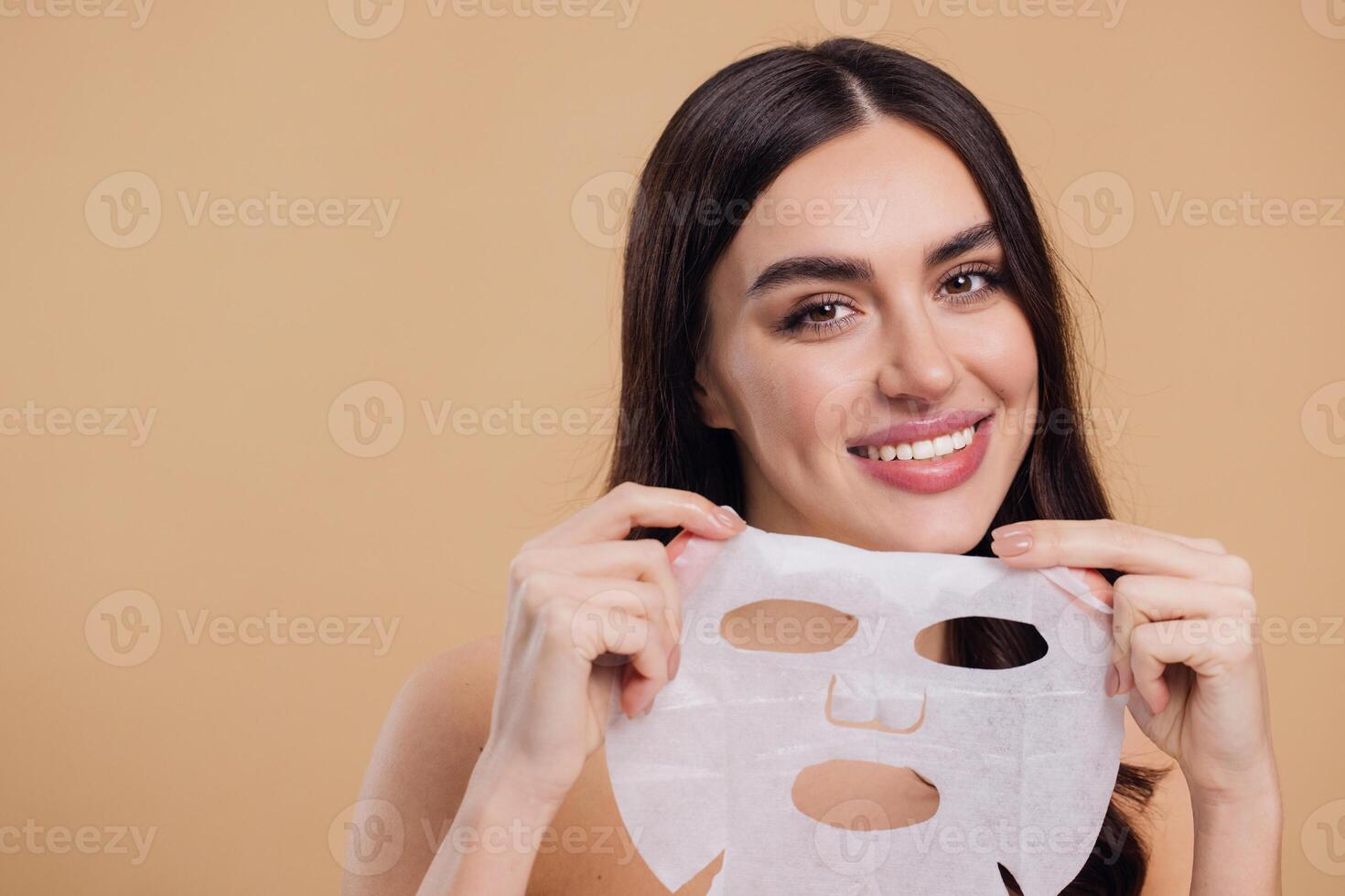 Beauty portrait of young woman with facial mask looking at camera over beige background photo