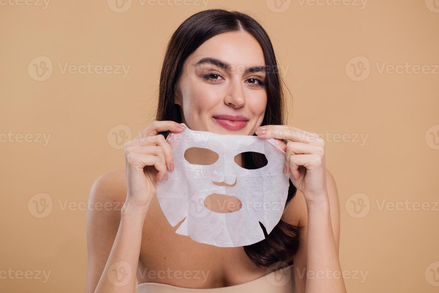 Beauty portrait of young woman with facial mask looking at camera over beige background photo