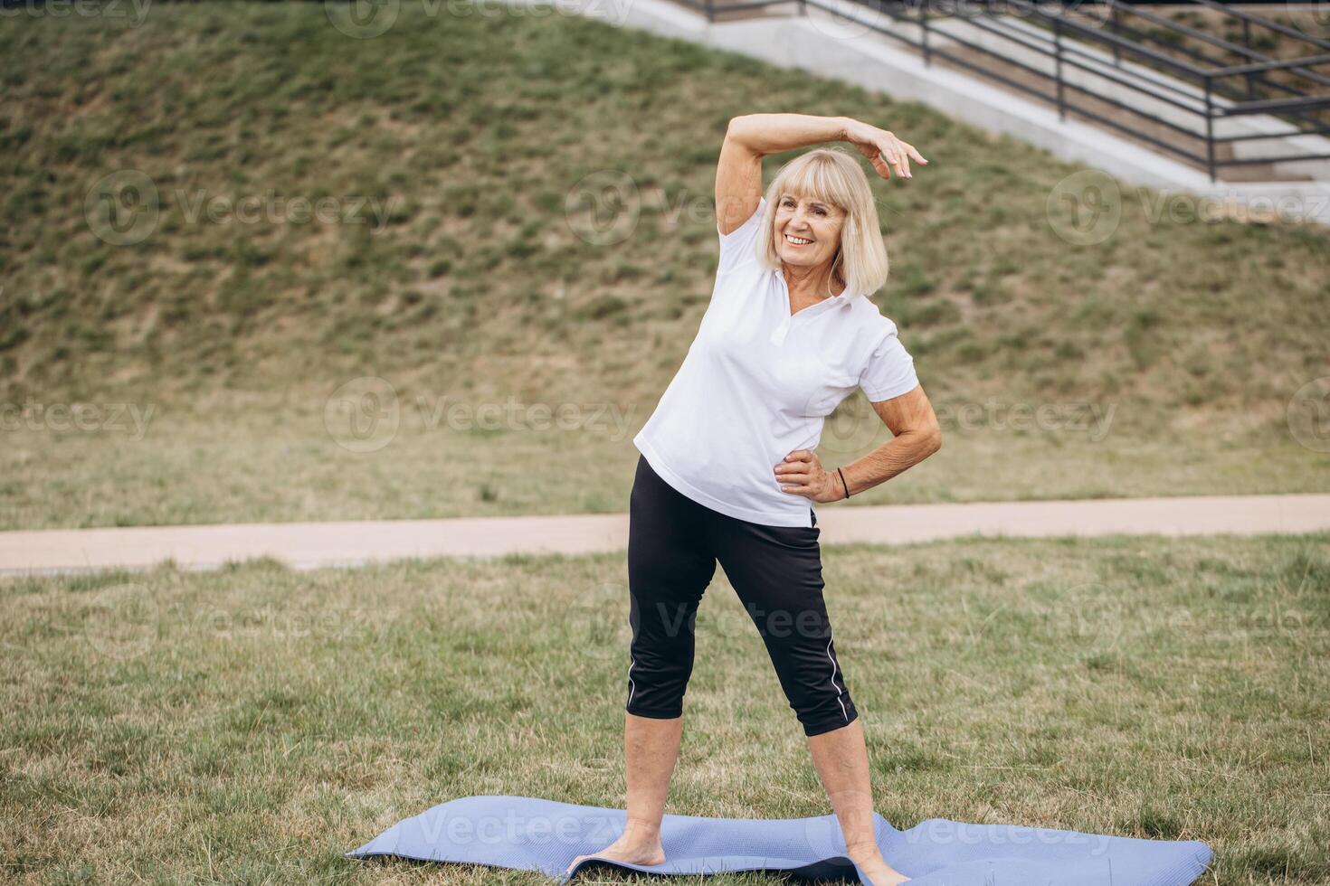Senior Woman Exercising Outdoors on Yoga Mat, Performing Stretching Routine in Park photo