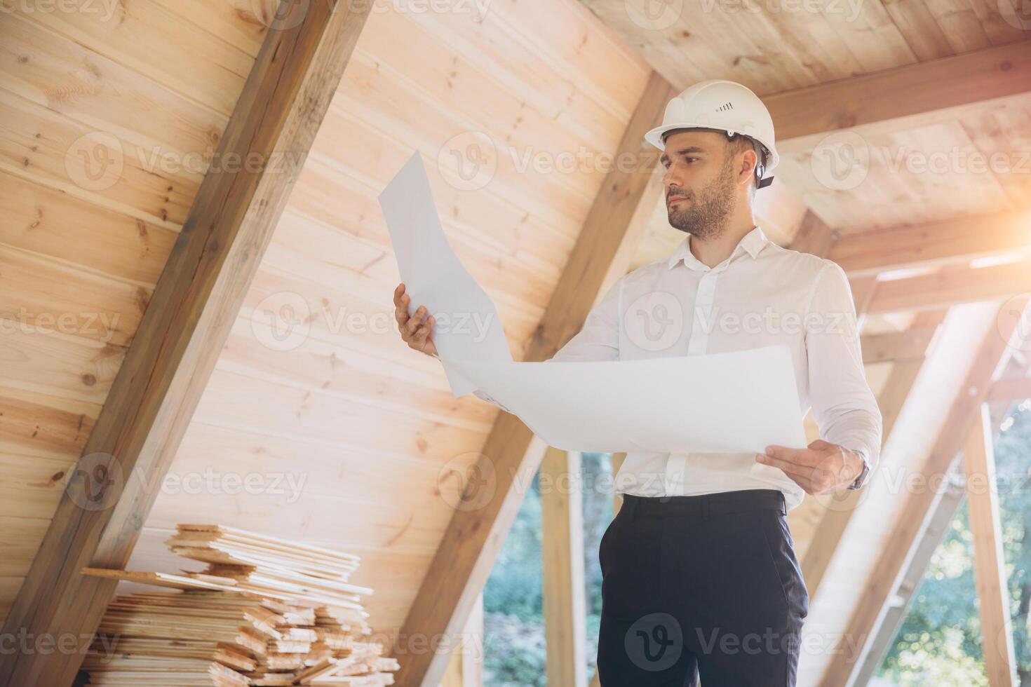 Architect wooden buildings. Man stands under frame wooden house. Man architect studies documentation or blueprints. Guy in protective helmet at construction site. Architect under timber framed house photo