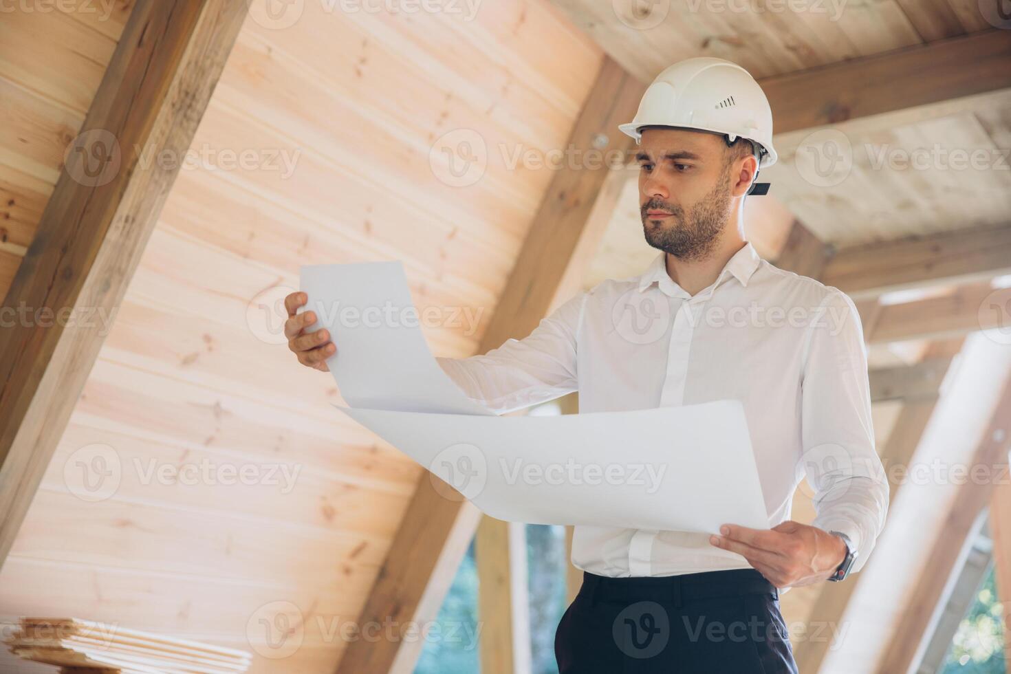 Architect wooden buildings. Man stands under frame wooden house. Man architect studies documentation or blueprints. Guy in protective helmet at construction site. Architect under timber framed house photo