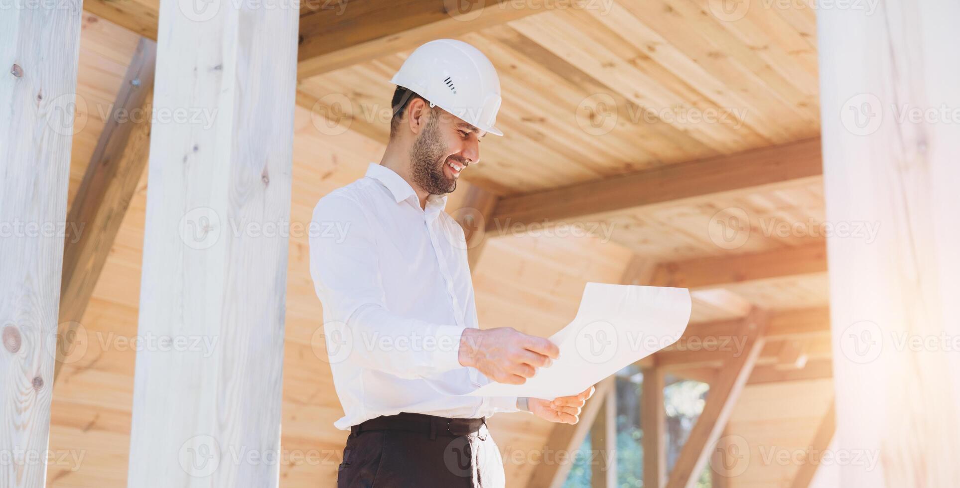 Construction engineer in white helmet. Developer with construction documentation on the background of wooden modular building under construction. Portrait of smiling architect near eco friendly house. photo