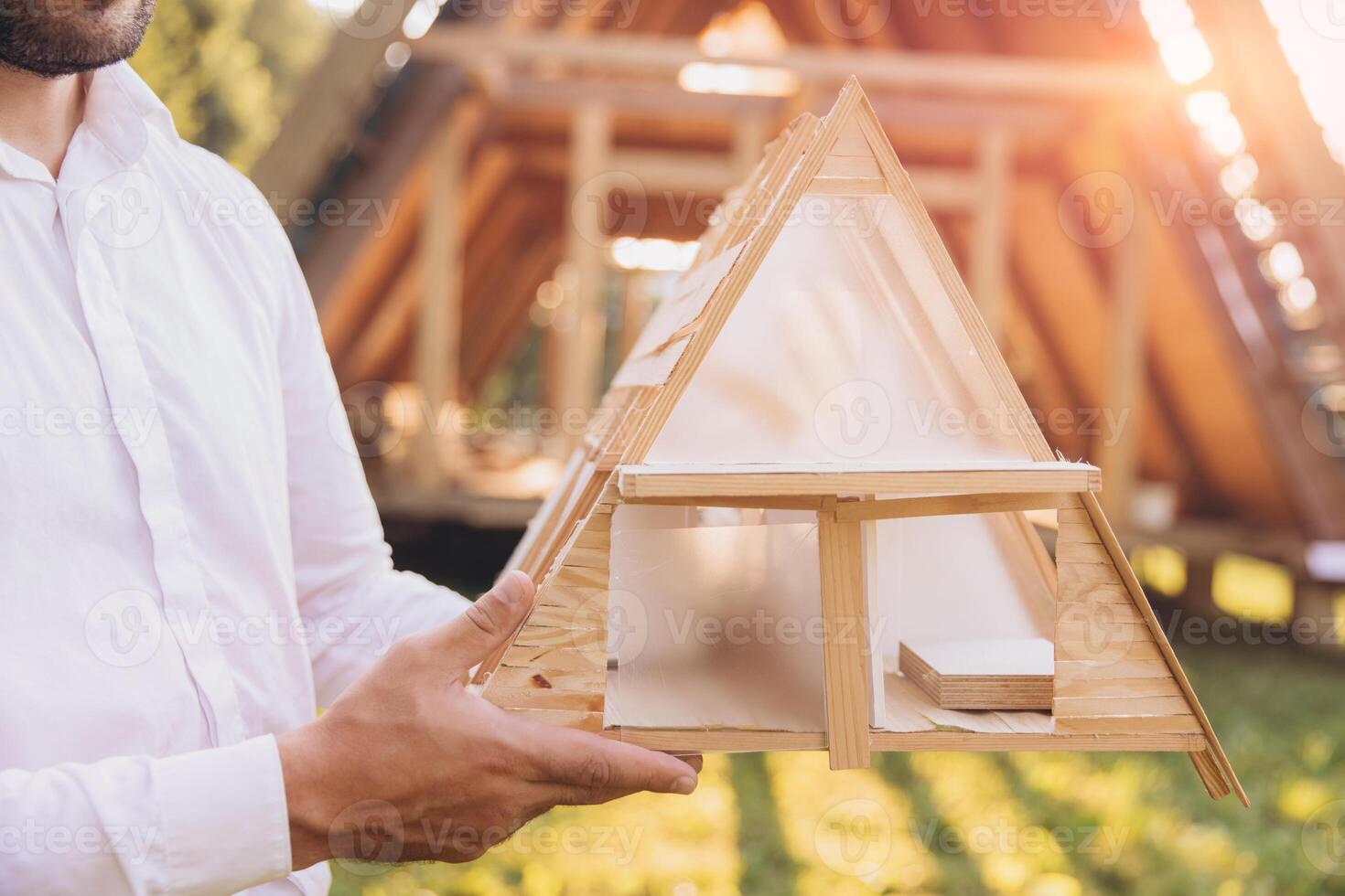 Man builder. Contractor for construction company. Architect in unfinished building. Builder in wooden modular house under construction. Close-up male engineer holding a model of miniature wooden house photo
