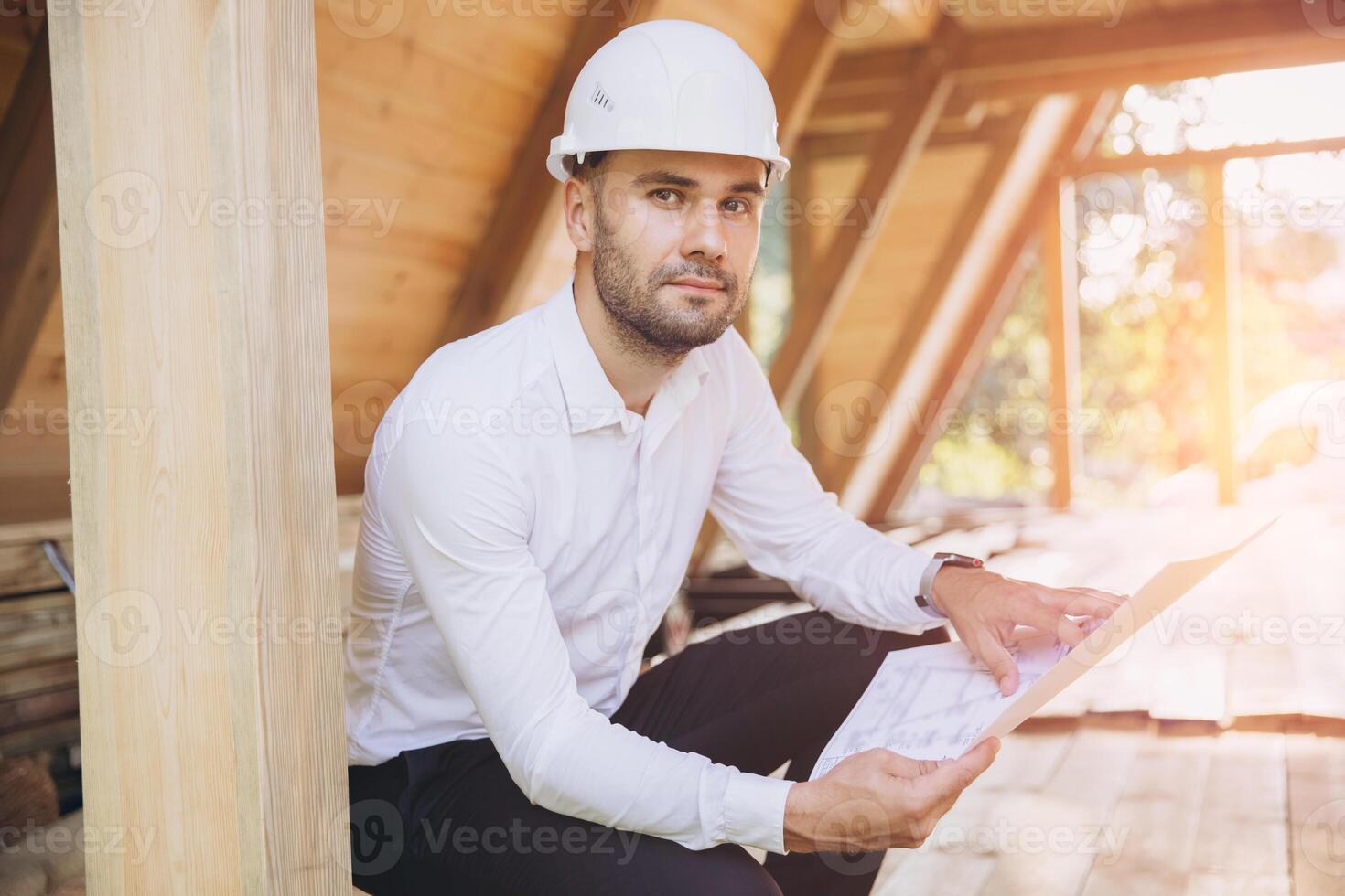 Architect wooden buildings. Man stands under frame wooden house. Man architect studies documentation or blueprints. Guy in protective helmet at construction site. Architect under timber framed house photo