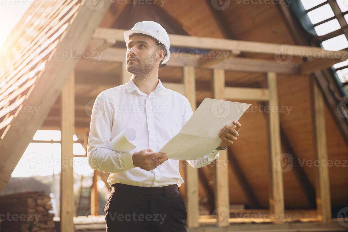 Architect wooden buildings. Man stands under frame wooden house. Man architect studies documentation or blueprints. Guy in protective helmet at construction site. Architect under timber framed house photo
